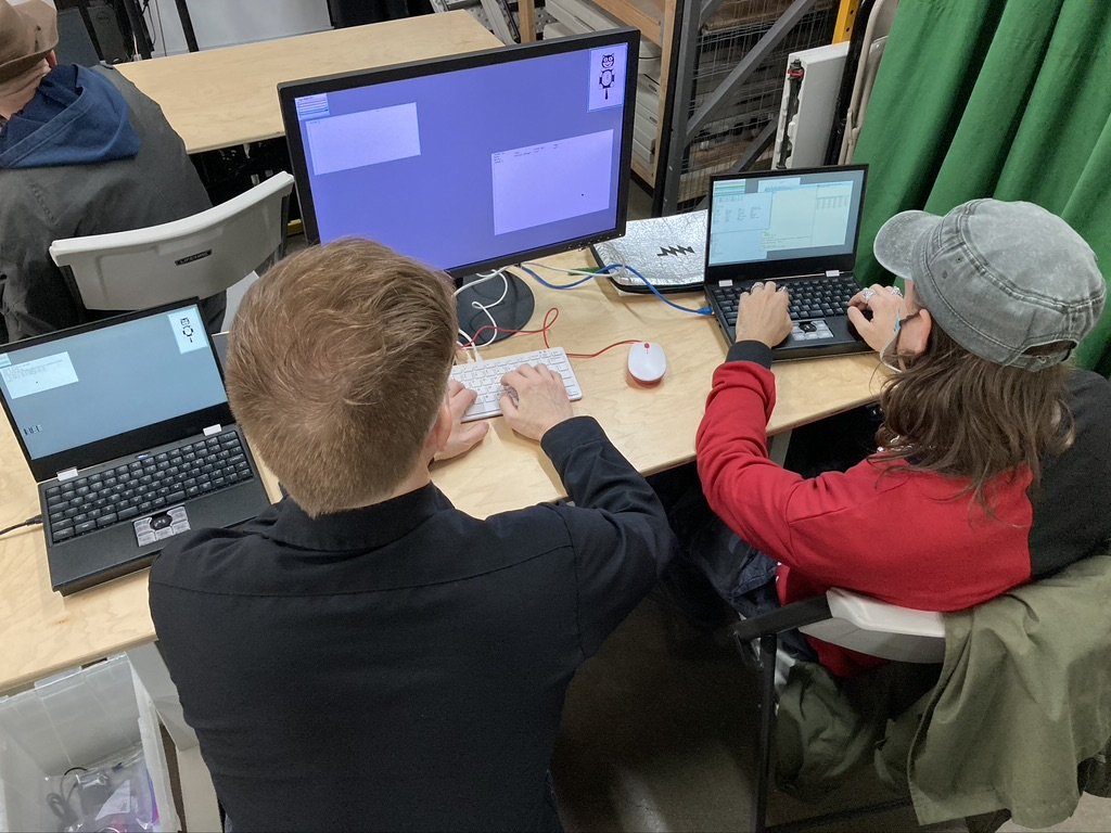 Two people sit at a desk with two MNT Reform laptops and a small monitor powered by a Raspberry Pi 400, all running 9front