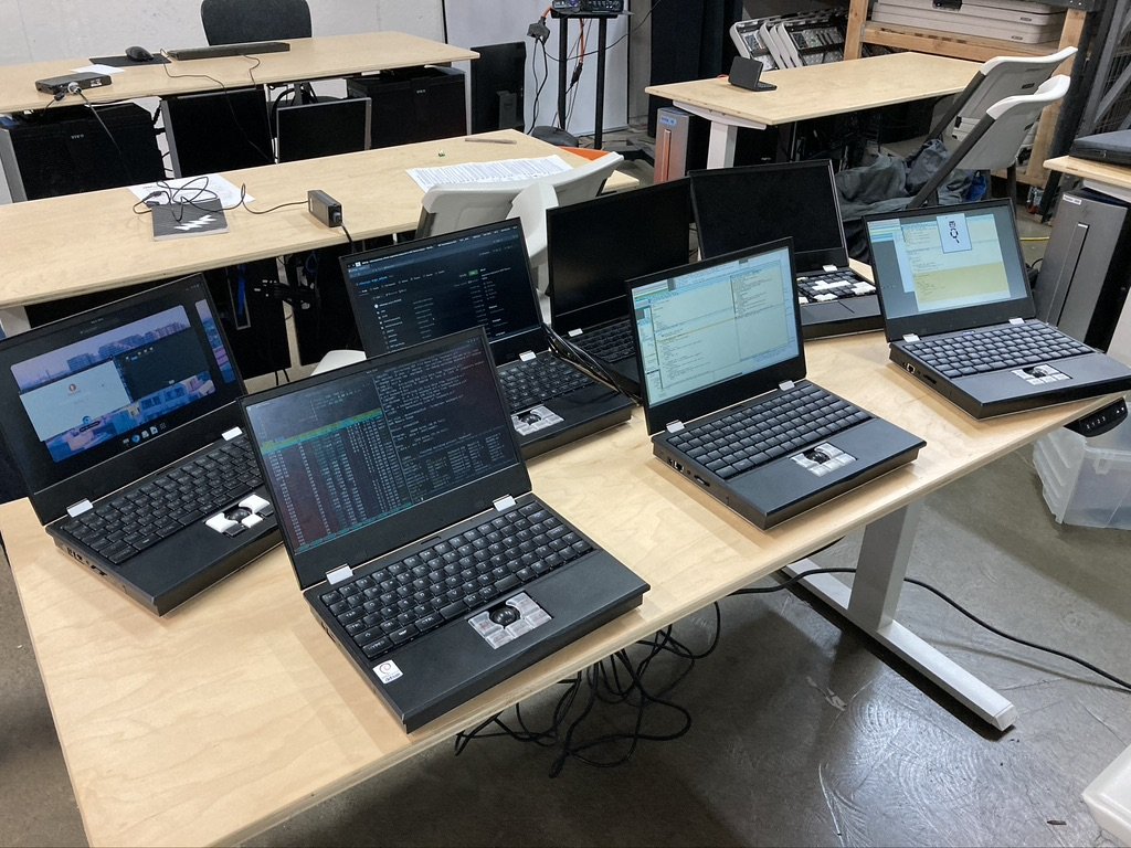 Seven MNT Reform laptops on a table together in a hackerspace