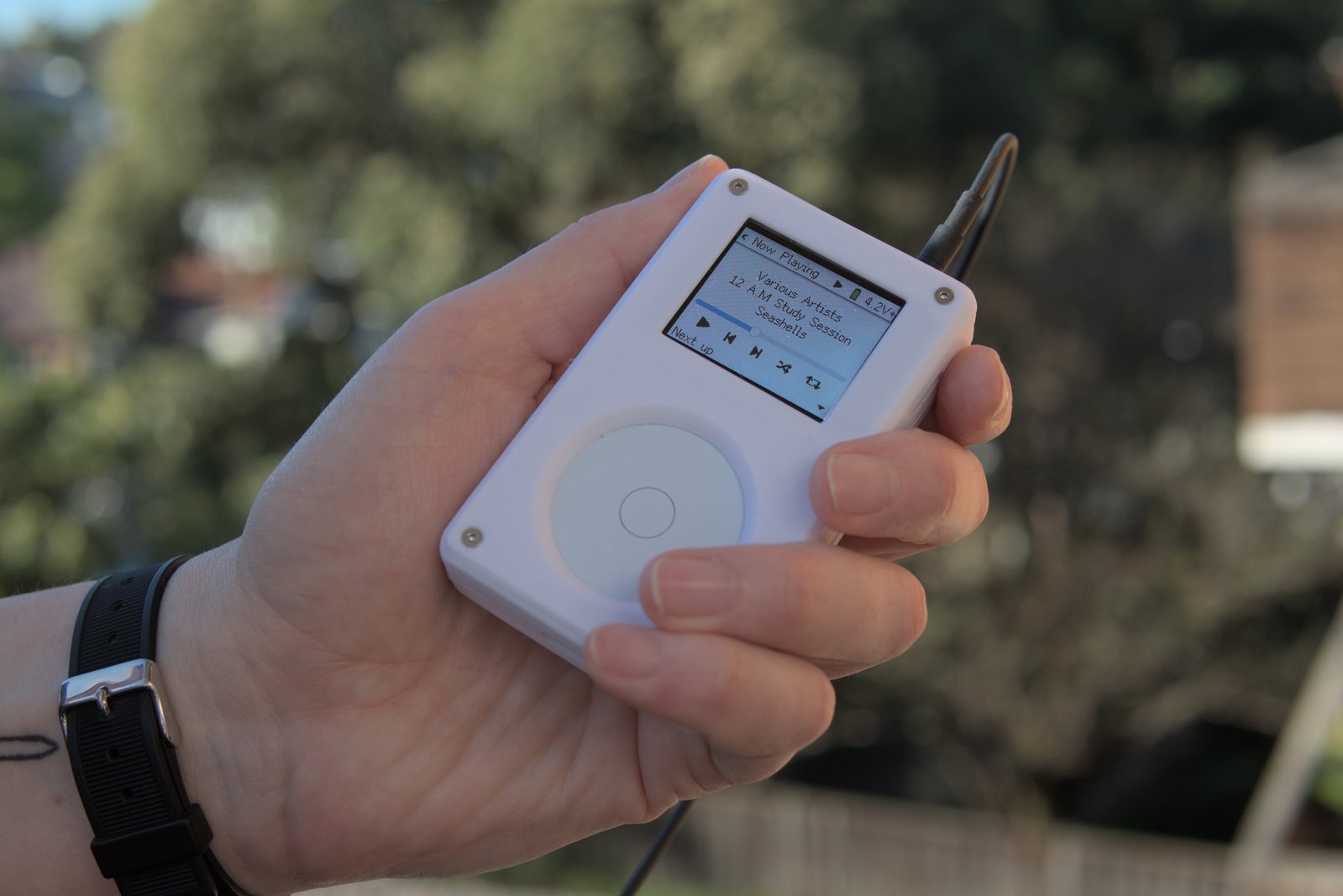 It's a Tangara music player! It's a little rectangular device being held in someone's hand. The front has a screen showing a 'Now Playing' screen, and a circular white touchwheel.