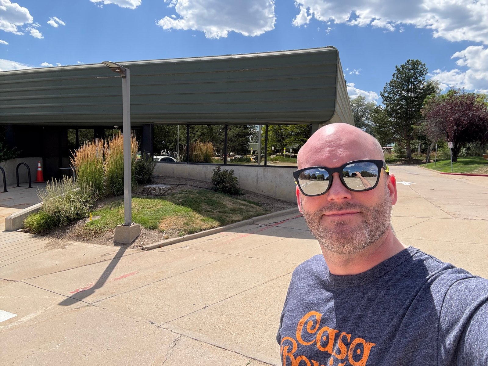 Selfie of me, a middle aged bald man with a stubbly beard, wearing mirrored sunglasses. I’m standing in front of a nondescript one story office building. It’s sunny and the skies are partly cloudy.