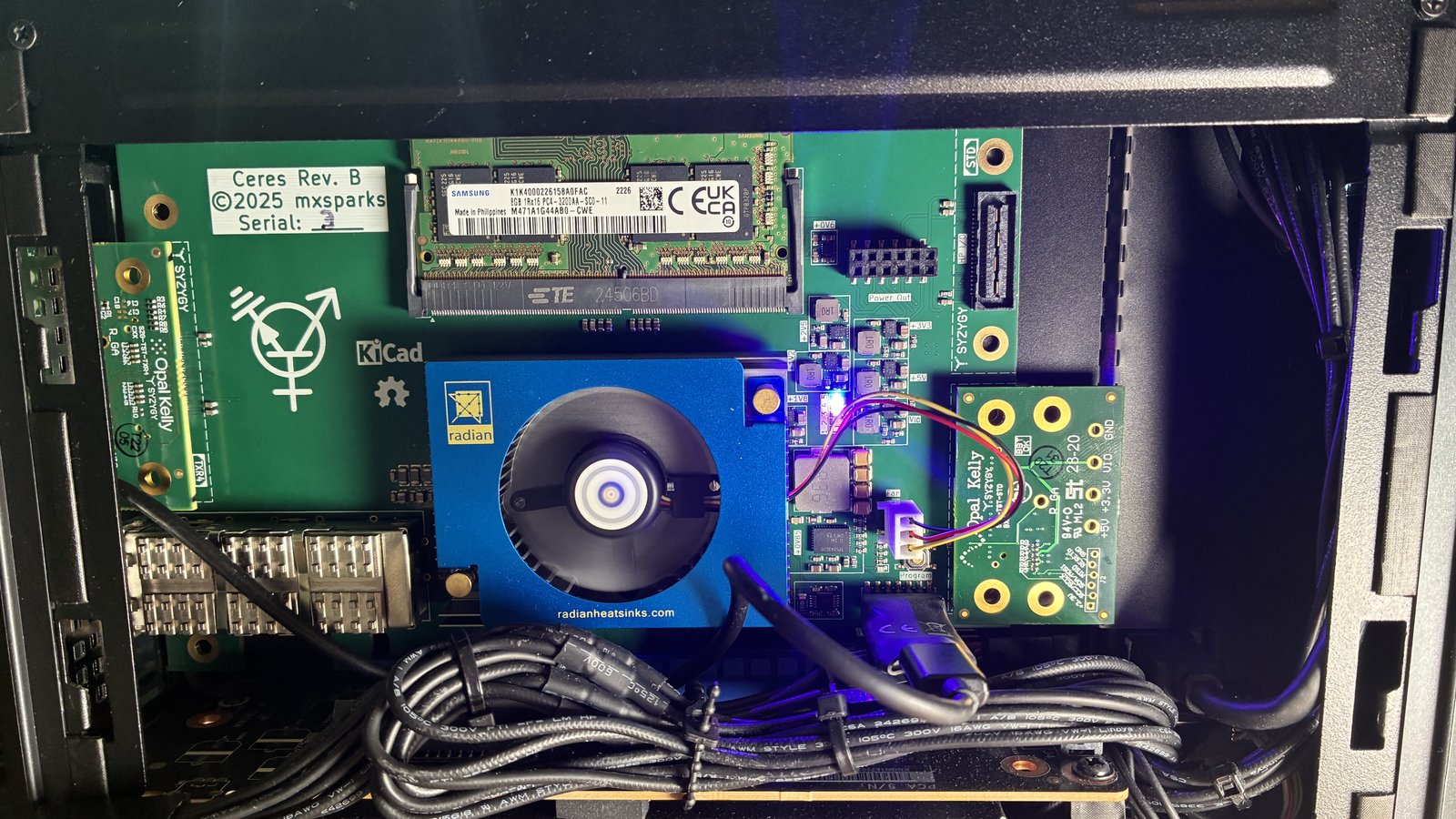 The same PCI Express card, now installed in a small PC chassis and with a DDR4 SODIMM and two SYZYGY test cards inserted. LEDs on the board are glowing blue, pink, and white.